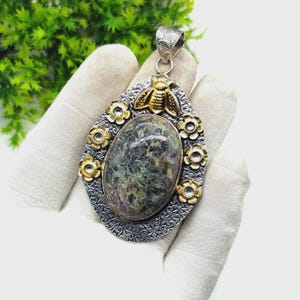 Pode incluir: Um pingente em forma de gota com uma grande pedra oval de tons verdes e roxos mesclados no centro. A moldura é de metal prateado texturizado, decorada com pequenas flores douradas e uma abelha dourada no topo, com uma argola prateada.