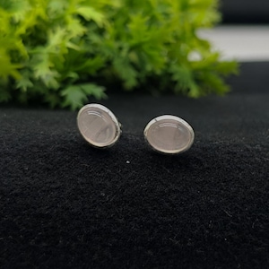 Op de afbeelding: Een paar oorknopjes met gladde, ovaalvormige lichtroze stenen in gepolijste zilverkleurige metalen zettingen. De oorbellen hebben een eenvoudig, afgerond ontwerp met een zachte, doorschijnende uitstraling en een lichte blushkleur.