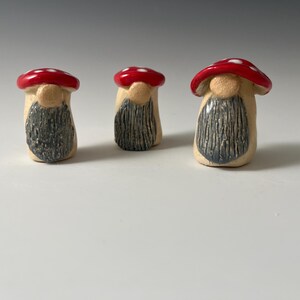 May include: Three ceramic mushroom figurines with red caps and white polka dots. The mushroom caps are glossy, and the bodies are beige with gray beards. The figurines are arranged on a white surface.