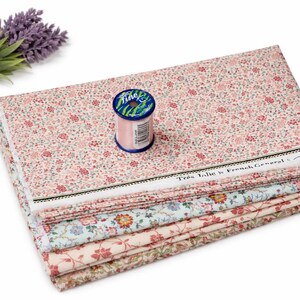 Könnte beinhalten: Ein Stapel Stoffe mit Blumenmustern in verschiedenen Farben, darunter Rosa, Blau und Creme, mit einer Spule rosa Garn und einem Lavendelzweig. Der Stoff trägt den Text "Très Jolie by French General".