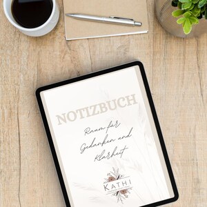 May include: A digital tablet with the word "NOTIZBUCH" and the text "Raum für Gedanken und Klarheit" is on a wooden surface. A cup of coffee, a notepad, a pen, and a small plant are also on the surface.