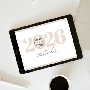 May include: A digital tablet displays a calendar design for the year 2026, with the word "Kalender" below. The design features the name "Kathi" and floral elements. A laptop, pen, and coffee cup are also visible.