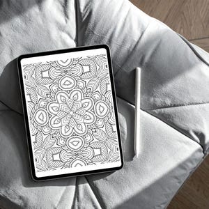 May include: A digital tablet displaying a black and white mandala design, with a white stylus resting beside it. The tablet has a black border and is placed on a gray, textured surface. The mandala features intricate floral and geometric patterns.