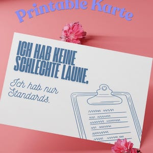 May include: A white printable card with blue text that reads "ICH HAB KEINE SCHLECHTE LAUNE. Ich hab nur Standards." A blue line drawing of a clipboard is on the right. Pink flowers are at the top and bottom.