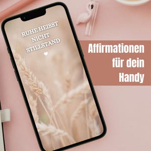 以下が含まれることがあります： スマートフォンには、小麦畑の画像の上に「RUHE HEISST NICHT STILLSTAND」というテキストが表示されています。「Affirmationen für dein Handy」という言葉も表示されています。ピンク色の背景には、コーヒーカップ、フラミンゴの置物、マウスがあります。