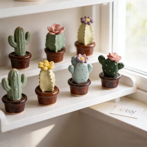 Puede incluir: Colección de figuras de cactus de cerámica en pequeñas macetas marrones, cada una con una flor de diferente color en la parte superior. Los cactus son de varios tonos de verde, amarillo y azul. Las figuras se exhiben en una estantería blanca.