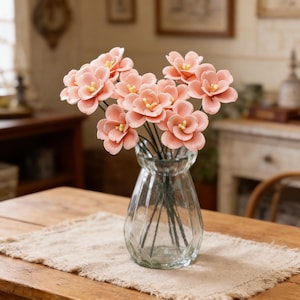 May include: A clear glass vase holds a bouquet of peach-colored flowers with yellow centers. The flowers are arranged on a wooden table with a beige linen runner. The vase has a ribbed design.