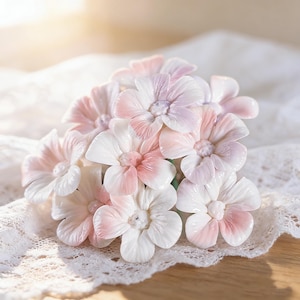 May include: A cluster of delicate, handcrafted flowers with white, pink, and lavender petals. The flowers are arranged on a white lace surface, creating a soft and elegant aesthetic. The petals have a glossy finish, and the overall design is detailed.