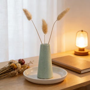 May include: A light green ceramic vase with a speckled design, holding three dried, fluffy stems. The vase sits on a white, round plate. A small lamp and a closed notebook are in the background.