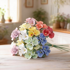 May include: A vibrant bouquet of ceramic flowers in various colours including pink, yellow, orange, red, blue, white, and green. The flowers are tied with green stems and twine, displayed on a light wooden surface.