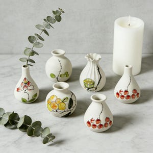 Könnte beinhalten: Eine Sammlung kleiner, weißer Keramikvasen mit handgemalten Blumen- und Fruchtdesigns. Die Vasen variieren in Form und Größe, einige haben einen gerüschten Rand. Eine weiße Säulenkerze und Eukalyptuszweige ergänzen das Arrangement.