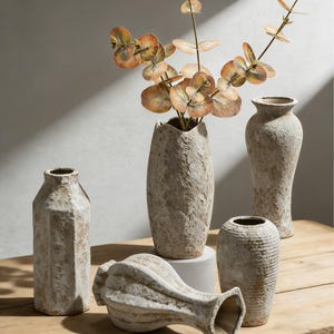 Puede incluir: Una colección de jarrones decorativos de varias formas y tamaños. Los jarrones tienen un acabado texturizado blanquecino, algunos con aspecto envejecido. Un jarrón contiene ramas de eucalipto artificiales con hojas marrones y verdes. Los jarrones están colocados sobre una superficie de madera.