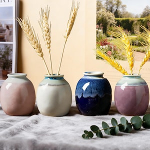 May include: Four ceramic vases in shades of pink, white, blue, and purple, each with a unique glaze. The vases hold dried wheat stalks. The vases are arranged on a gray, textured surface.