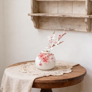Puede incluir: Un jarrón de cerámica blanca con detalles florales rosas y una rama de flores blancas y rosas. El jarrón está sobre una mesa de madera con un tapete de encaje y un camino de mesa de lino. Un estante de madera rústica en el fondo.