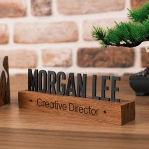May include: A desk name plate featuring the black text MORGAN LEE in a bold sans-serif font mounted on a rectangular wooden base. The base is engraved with the text Creative Director and two small diamond symbols on the front face.