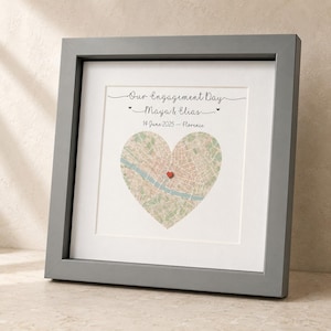 May include: A square gray frame containing a white mat and a print featuring the text Our Engagement Day, Maya & Elias, and 14 June 2025 - Florence. A heart-shaped map of a city is centered with a small red heart marker placed on the map.