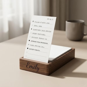 Puede incluir: Un portanotas de escritorio de madera con el nombre "Emily" grabado en la parte delantera. Un bloc de notas blanco con una lista de tareas se inserta en el soporte. La lista incluye elementos como "Follow up with Josh" y "Water Plants."