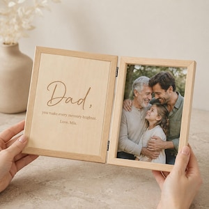 May include: A hinged light wood double photo frame. The left panel features the brown printed text: Dad, you make every memory brighter. Love, Mia. The right panel contains a color photograph of an older man, a younger man, and a young girl.