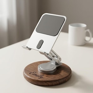 May include: A silver and gray adjustable phone stand with a dark gray pad, mounted on a round wooden base. The base is engraved with the name "Emily" in cursive. A white mug is in the background.