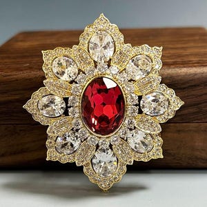 Puede incluir: Broche dorado con diseño de estrella. Presenta una gran gema roja ovalada en el centro, rodeada de cristales transparentes y brillantes. La broche está colocada sobre una caja de madera.