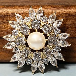 May include: A circular brooch featuring a central white pearl surrounded by multiple layers of clear, marquise-cut and round-cut cubic zirconia stones set in a gold-toned metal frame, arranged in a sunburst pattern.