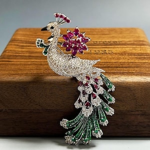 May include: A peacock-shaped brooch featuring a silver-toned body encrusted with clear crystals, a crest of red crystals, a floral accent of red marquise-cut crystals, and a long, flowing tail decorated with alternating red and green crystal accents.