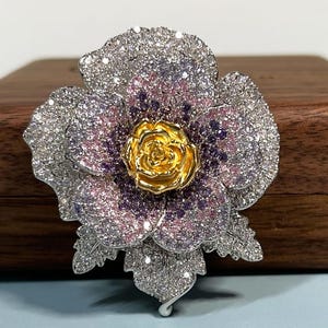 May include: A floral brooch featuring a central gold-toned rose surrounded by layered petals encrusted with white, pink, and purple crystals. The petals have a silver-toned metal base, and the entire piece is covered in sparkling pavé stones.