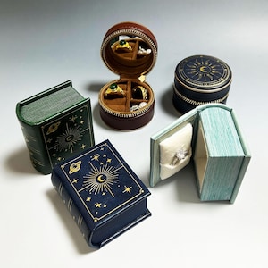 May include: An assortment of small, decorative jewelry boxes in various shapes and colors. There's a round, brown zippered case, a dark blue box, and two book-shaped boxes, one green and one blue. Each box features celestial designs in gold.