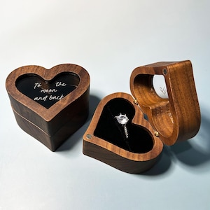 May include: Two heart-shaped wooden ring boxes with a dark brown finish. One box is closed with the text "To the moon and back" printed on the lid. The other box is open, revealing a black velvet interior holding a silver ring with a clear stone.