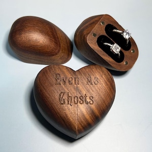 May include: A heart-shaped dark wood ring box with the text Even As Ghosts engraved on the front. The box is shown open with two silver rings with clear stones inside, and the detached lid is placed to the side of the main container.