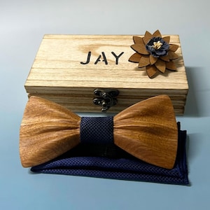 May include: A wooden bow tie with a navy blue centre and matching pocket square. The set includes a wooden box with the name "JAY" engraved on the lid, and a wooden flower lapel pin. The bow tie is made of light brown wood.