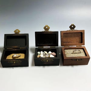 Può includere: Tre scatole portanelli in legno aperte con ferramenta in ottone. Una scatola contiene sabbia e un anello, un'altra piccole pietre e la terza un anello su tela di juta. Le scatole sono tinte in marrone scuro e tonalità legno naturale.