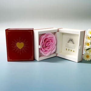 May include: A red book-shaped jewelry box with a gold heart design on the cover. Inside, a pink rose and a diamond ring are displayed. The ring is in a compartment with the name "LINA" in gold lettering.