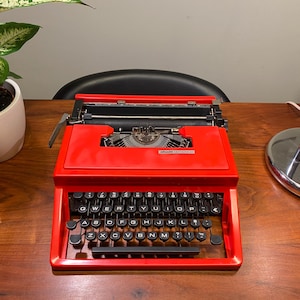 Peut inclure: Une machine à écrire rouge vif avec des touches noires est posée sur une surface en bois. La machine à écrire présente un design classique avec un levier de retour de chariot et un mécanisme d'alimentation du papier. Les touches sont disposées en QWERTY. Une plante en pot est en arrière-plan.