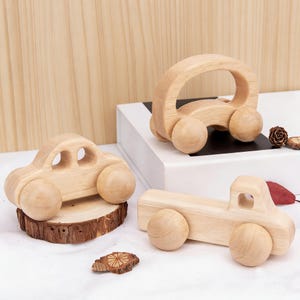 Może przedstawiać: Trzy drewniane zabawki: samochód, zaokrąglony samochód i ciężarówka. Zabawki wykonane są z jasnego drewna i mają zaokrąglone koła. Samochód stoi na małym kawałku drewna. Tło to jasne drewno.