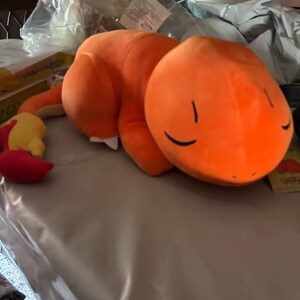 May include: An orange plush toy in the shape of a cartoon character, lying on a table. The toy has a rounded head with closed eyes and a long tail with a yellow and red tip. The plush is made of soft material.