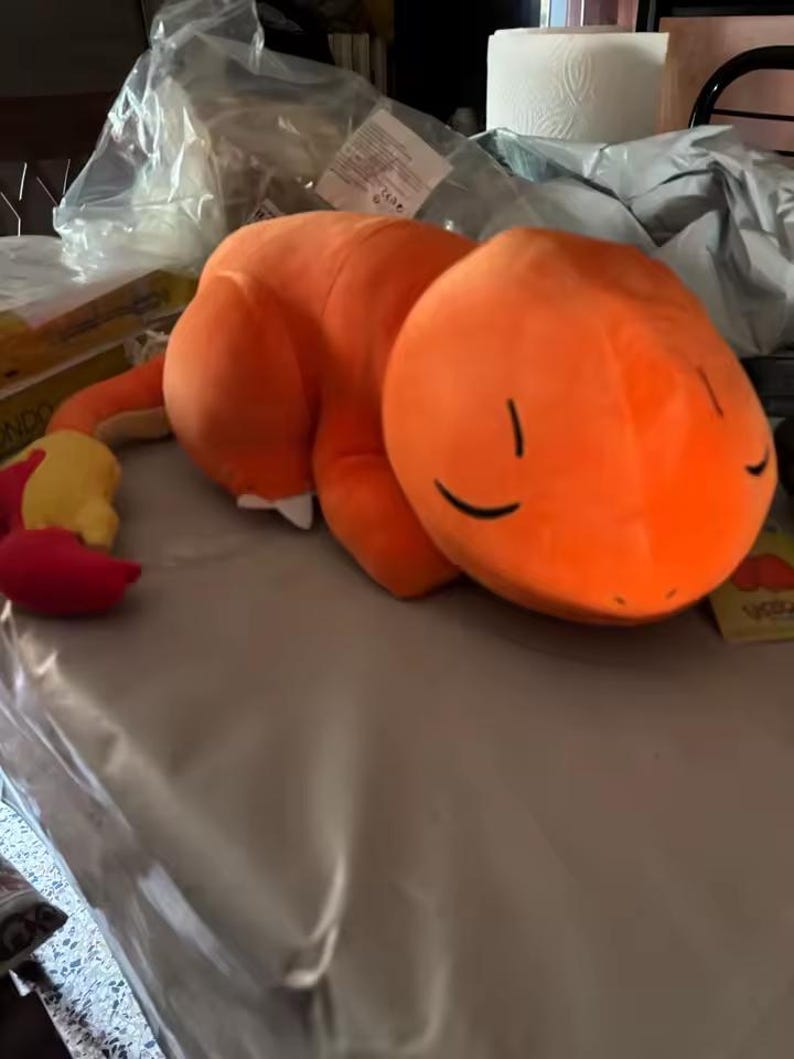 May include: An orange plush toy in the shape of a cartoon character, lying on a table. The toy has a rounded head with closed eyes and a long tail with a yellow and red tip. The plush is made of soft material.