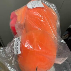 May include: An orange plush toy, packaged in clear plastic. The toy has a rounded shape with a small, black line drawn on the bottom. A tag is visible on the top of the package.