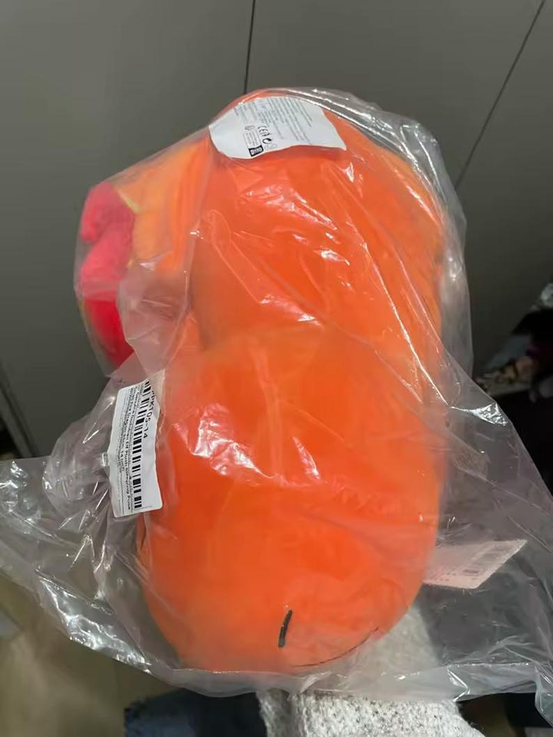 May include: An orange plush toy, packaged in clear plastic. The toy has a rounded shape with a small, black line drawn on the bottom. A tag is visible on the top of the package.