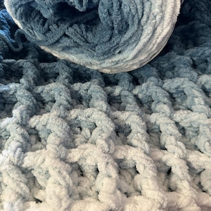 May include: A skein of soft, plush chenille yarn featuring an ombre color transition from deep navy blue to light sky blue and white. The yarn is worked into a waffle stitch pattern, creating a textured, grid-like fabric with a thick, fuzzy feel.