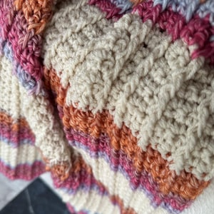 May include: A crocheted textile featuring a repeating striped pattern in cream, burnt orange, magenta, and soft lavender. The fabric displays a textured, ribbed stitch construction with vertical columns of raised yarn loops throughout the surface.