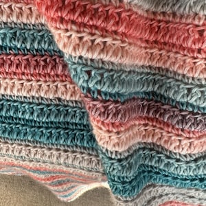 May include: A crocheted blanket featuring horizontal stripes in alternating shades of coral pink, muted teal, and light gray. The textile displays a consistent double crochet stitch pattern, creating a textured, rippled surface with soft, folded fabric.