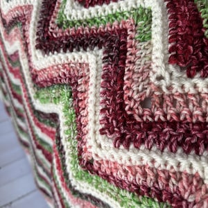 May include: A crocheted blanket featuring a repeating chevron pattern in shades of cream, light pink, deep burgundy, and various tones of green. The textile displays a textured stitch construction with distinct zigzag lines running vertically.
