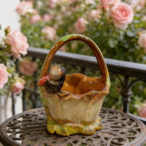 Puede incluir: Una cesta de cerámica con asa marrón y figura de pájaro. La cesta es de color beige con detalles verdes y marrones, y está sobre una mesa de metal decorativa. Rosas rosas en el fondo.