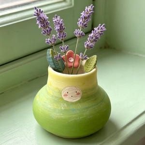 May include: A small, hand-painted ceramic vase in shades of green and yellow, adorned with a whimsical face. The vase holds sprigs of purple lavender, a pink flower, and green leaves. The vase is sitting on a green surface.