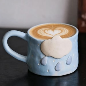 May include: A light blue ceramic coffee mug with a cloud and raindrop design. The mug is filled with a latte, topped with a heart-shaped design. The mug has a curved handle.