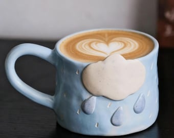 Peut inclure: Tasse à café en céramique bleu clair avec un motif de nuage et de gouttes de pluie. La tasse est remplie d'un latte, surmonté d'un motif en forme de cœur. La tasse a une anse incurvée.