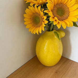 Könnte beinhalten: Eine leuchtend gelbe Keramikvase in Zitronenform, die mehrere Sonnenblumen enthält. Die Vase hat eine glänzende Oberfläche und ein grünes Blattdetail. Die Sonnenblumen haben leuchtend gelbe Blütenblätter und braune Zentren.