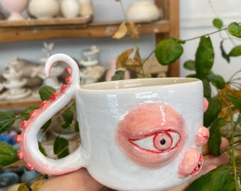Peut inclure: Tasse en céramique blanche avec un design unique représentant un œil rose et des tentacules. La poignée a la forme d'un tentacule avec des accents rouges. La tasse a une surface texturée et est probablement faite à la main.