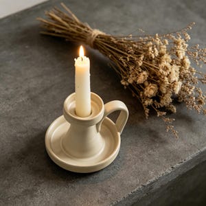 May include: A lit white candle in a cream-coloured ceramic candlestick holder with a handle. A bundle of dried flowers is behind the candle holder. The candle has a bright flame.
